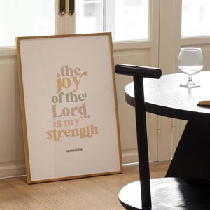 May include: A framed print with the text "the joy of the Lord is my strength" in a warm color palette. The print is displayed in a room with a black chair, a round table, and a glass of water.