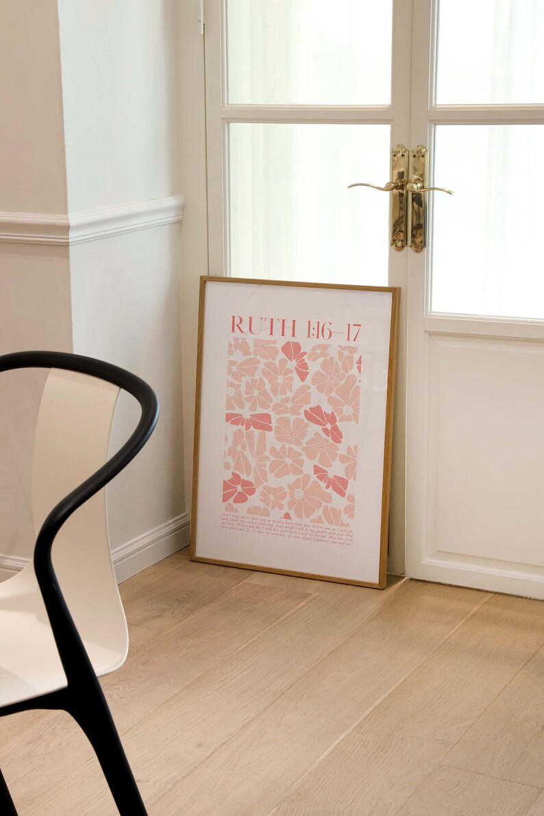 May include: A framed art print featuring a floral pattern in shades of pink and white, with the text "RUTH 1:16-17" at the top. The print is leaning against a white door with gold hardware, on a light wood floor.