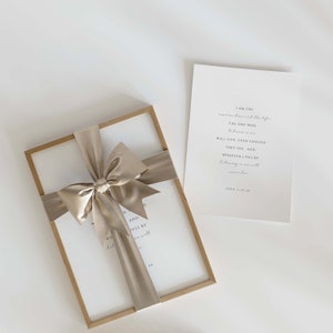May include: A framed print with a beige ribbon tied in a bow. The print features a quote from the Bible, John 11:25-26, "I am the resurrection and the life. The one who believes in me will live, even though they die; and whoever lives by believing in me will never die."