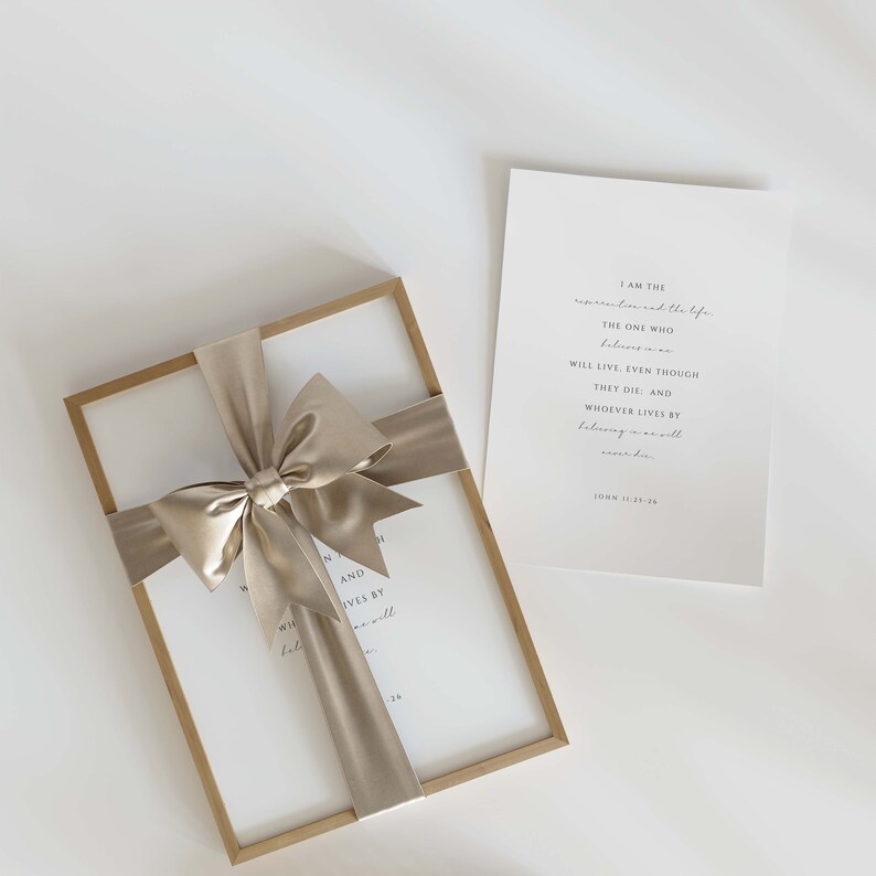 May include: A framed print with a beige ribbon tied in a bow. The print features a quote from the Bible, John 11:25-26, "I am the resurrection and the life. The one who believes in me will live, even though they die; and whoever lives by believing in me will never die."