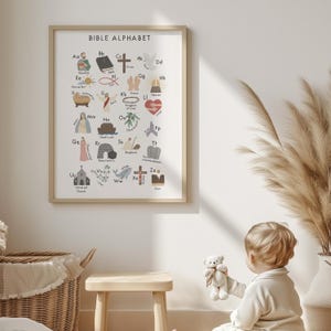 May include: A framed print titled "BIBLE ALPHABET" features colorful illustrations for each letter. The artwork includes religious symbols like a cross, dove, and heart, alongside related words. The print is in a light wood frame.