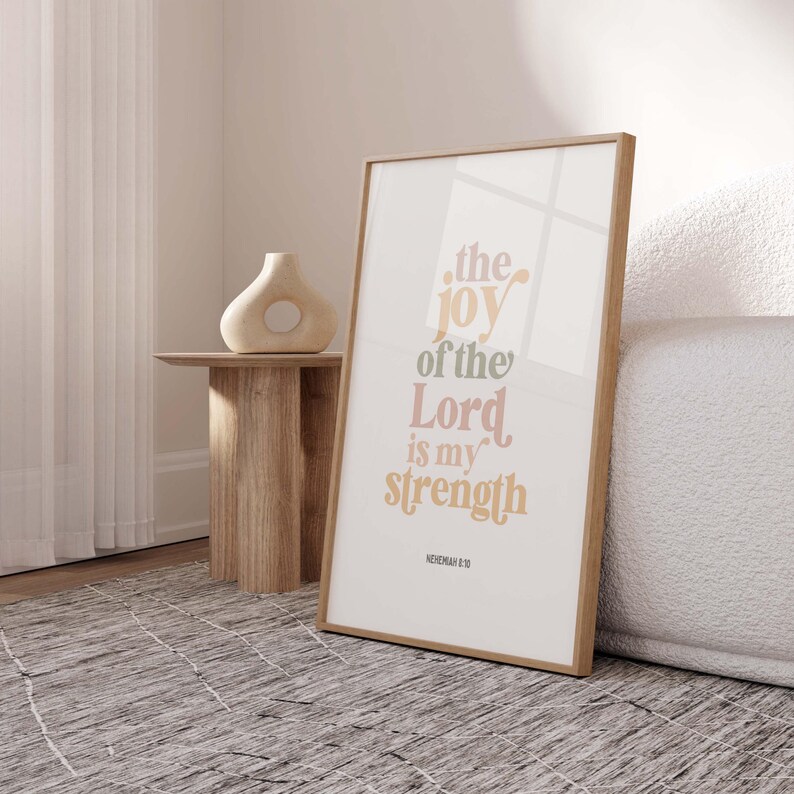May include: A framed print with the text "the joy of the Lord is my strength" in a stylish font. The print has a light wood frame and is displayed on a neutral-toned background. A decorative vase sits on a small wooden side table.