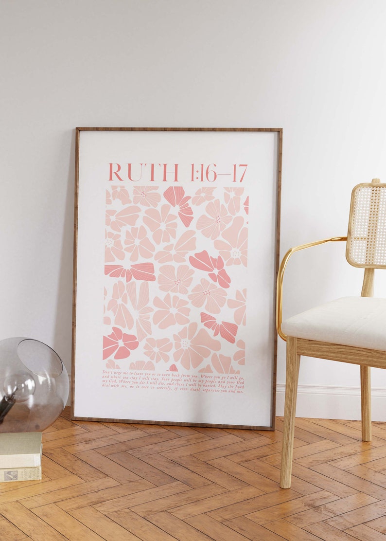 May include: A framed art print featuring a floral design in shades of pink. The print includes the text "RUTH 1:16-17" at the top and a passage below. The artwork is displayed on a white wall, next to a chair.