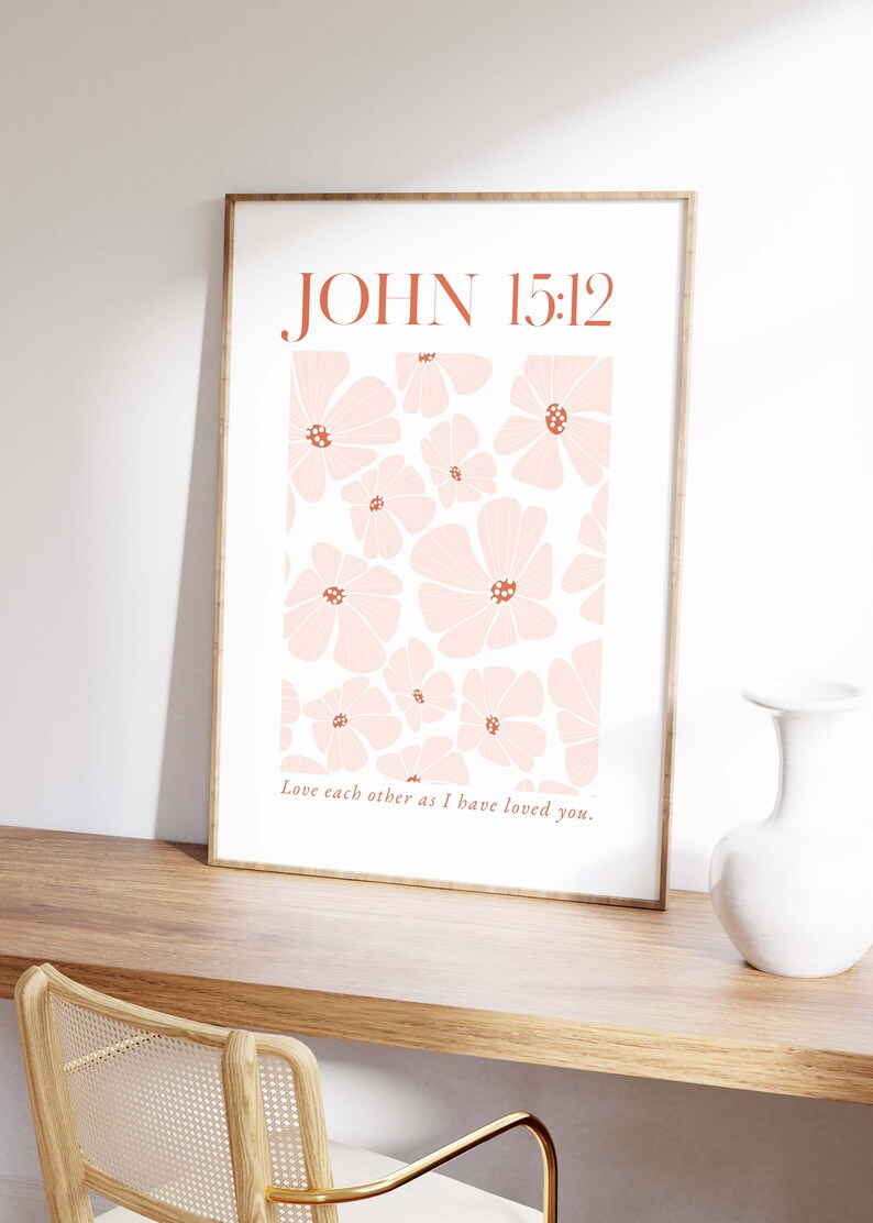 May include: A framed print with a white background and a pattern of pink flowers with red centers. The text "JOHN 15:12" is in brown at the top of the print. The text "Love each other as I have loved you." is in brown at the bottom of the print.