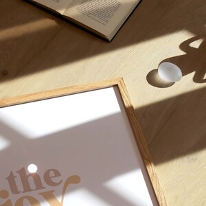 May include: A framed print with the words "the joy of the" in a retro font, in shades of peach, beige, and green. An open book and a white egg-shaped object are on a wooden surface.