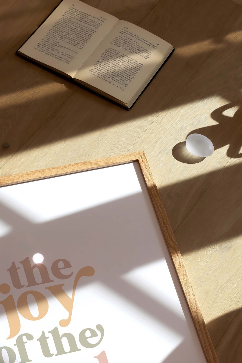 May include: A framed print with the words "the joy of the" in a retro font, in shades of peach, beige, and green. An open book and a white egg-shaped object are on a wooden surface.