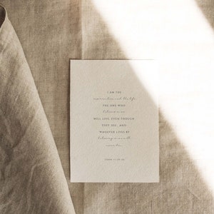 May include: A white card with a quote from the Bible, John 11:25-26, printed in black ink. The quote reads: "I am the resurrection and the life. The one who believes in me will live, even though they die. And whoever lives by believing in me will never die."