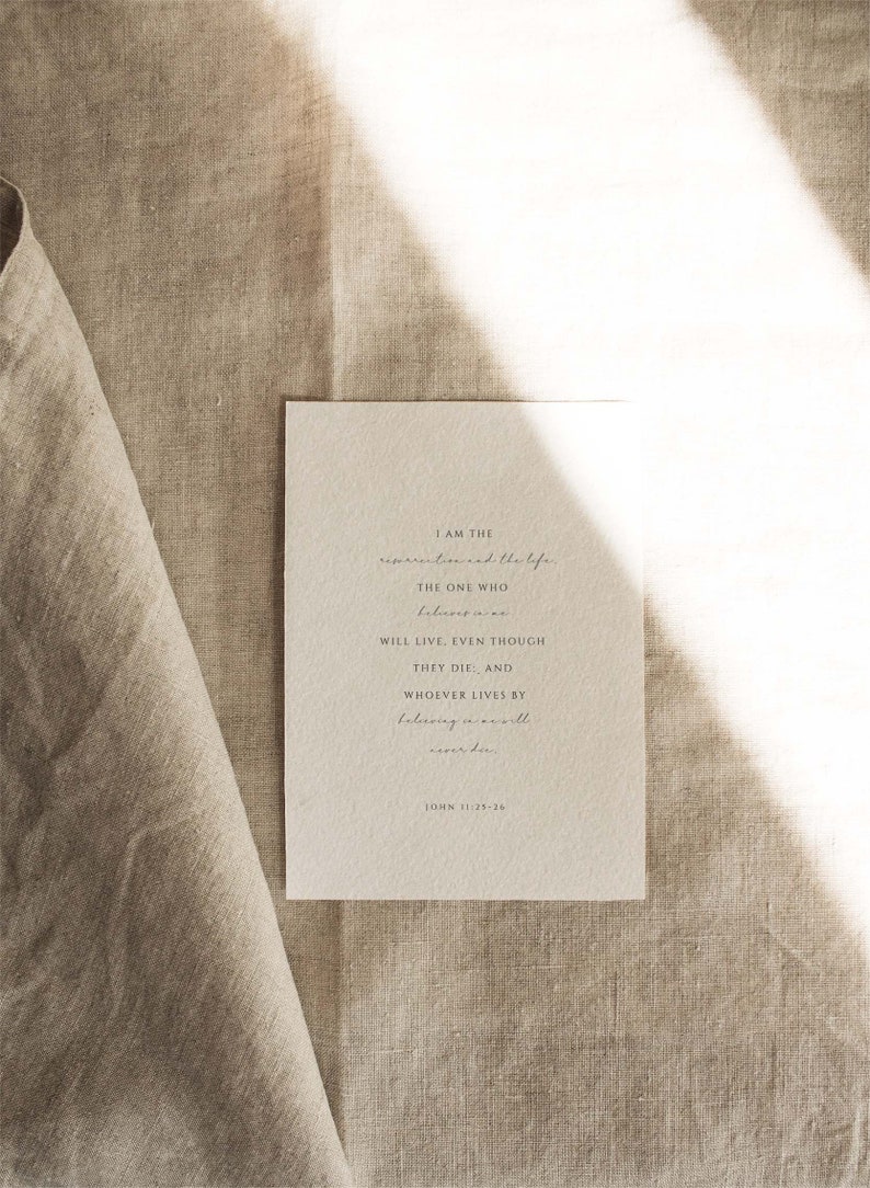 May include: A white card with a quote from the Bible, John 11:25-26, printed in black ink. The quote reads: "I am the resurrection and the life. The one who believes in me will live, even though they die. And whoever lives by believing in me will never die."