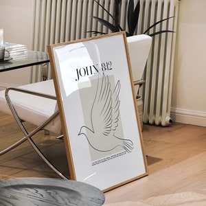 May include: A framed print with a minimalist design featuring a dove in flight and the text "JOHN 8:12" in black. The dove is outlined in black against a light beige background. The frame is a light wood colour.