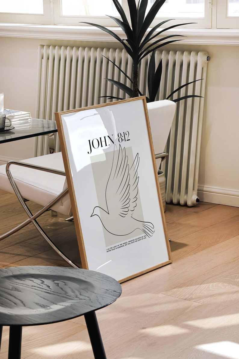 May include: A framed print with a minimalist design featuring a dove in flight and the text "JOHN 8:12" in black. The dove is outlined in black against a light beige background. The frame is a light wood colour.