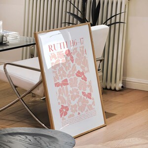 May include: A framed art print featuring the text "RUTH 1:16-17" in coral, with a floral design in shades of pink. The print is displayed in a light wood frame, leaning against a white chair in a well-lit room.