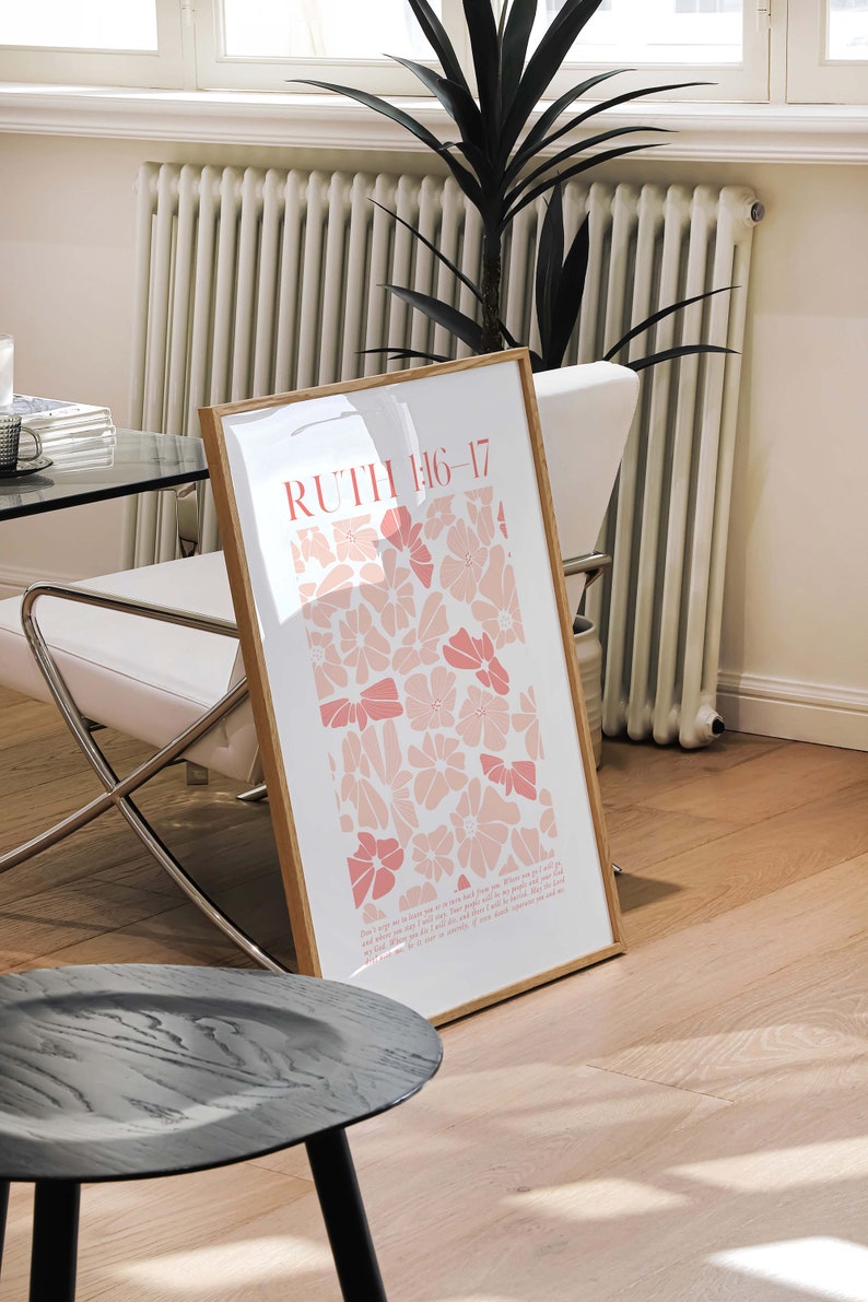 May include: A framed art print featuring the text "RUTH 1:16-17" in coral, with a floral design in shades of pink. The print is displayed in a light wood frame, leaning against a white chair in a well-lit room.