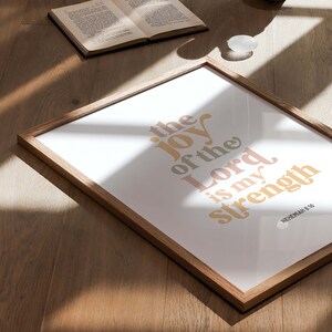 May include: A framed print with the text "the joy of the Lord is my strength" in a retro font. The print is in a wooden frame and is on a wooden surface. An open book and a vase are also on the surface.