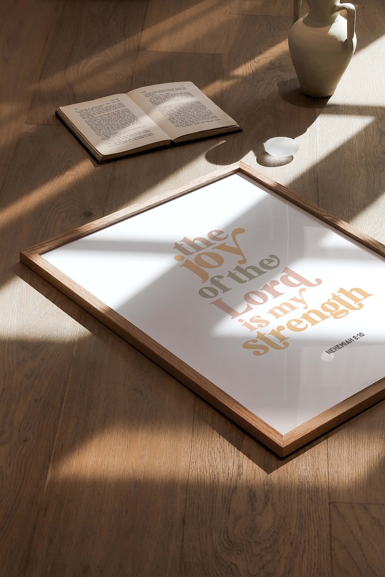 May include: A framed print with the text "the joy of the Lord is my strength" in a retro font. The print is in a wooden frame and is on a wooden surface. An open book and a vase are also on the surface.