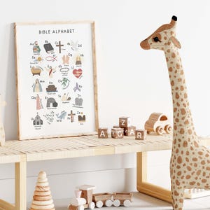 May include: A framed "Bible Alphabet" print with colorful illustrations and text, displayed on a light wood bench. A large stuffed giraffe with brown spots stands nearby, along with wooden toys and alphabet blocks.
