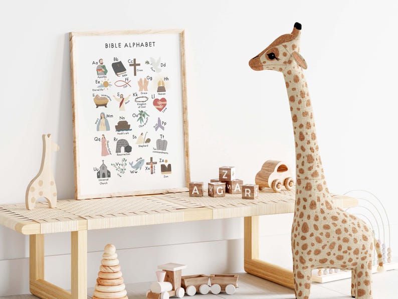 May include: A framed "Bible Alphabet" print with colorful illustrations and text, displayed on a light wood bench. A large stuffed giraffe with brown spots stands nearby, along with wooden toys and alphabet blocks.