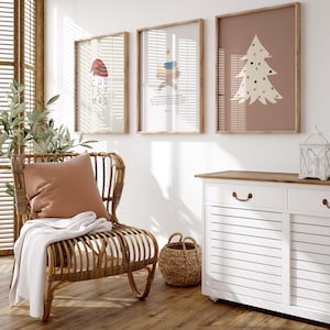 May include: Three framed Christmas-themed prints with a white background and brown frames. The prints feature a red Santa hat, a nativity scene, and a stylized Christmas tree with colorful ornaments. A white wicker chair with a brown cushion and a white throw blanket is in the foreground. A white cabinet with two drawers and a wooden top is in the background.