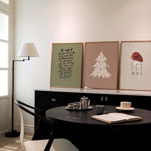 May include: Three framed prints on a wall, one with a green background and a black text quote, "The word became flesh and made his dwelling among us. We have seen his glory, the glory of the only Son, who came from the Father, full of grace and truth." The second print has a light brown background with a white stylized Christmas tree with colorful ornaments. The third print has a light pink background with a red Santa hat and the text "CHRISTMAS".