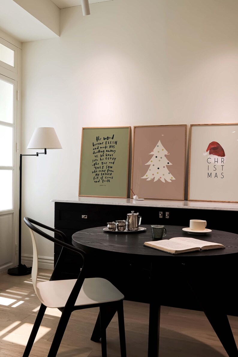 May include: Three framed prints on a wall, one with a green background and a black text quote, "The word became flesh and made his dwelling among us. We have seen his glory, the glory of the only Son, who came from the Father, full of grace and truth." The second print has a light brown background with a white stylized Christmas tree with colorful ornaments. The third print has a light pink background with a red Santa hat and the text "CHRISTMAS".