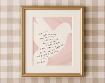 May include: A framed print with a gold-colored frame. The artwork features a quote in black text on a cream-colored background, with a pink abstract shape. The quote is from Romans 15:13, offering a message of hope and faith.