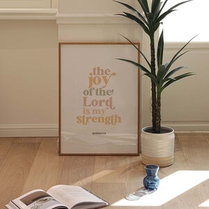 May include: A framed print with the text "the joy of the Lord is my strength" in a warm color palette. A potted plant, an open book, and a small blue vase are also visible on the wooden floor.