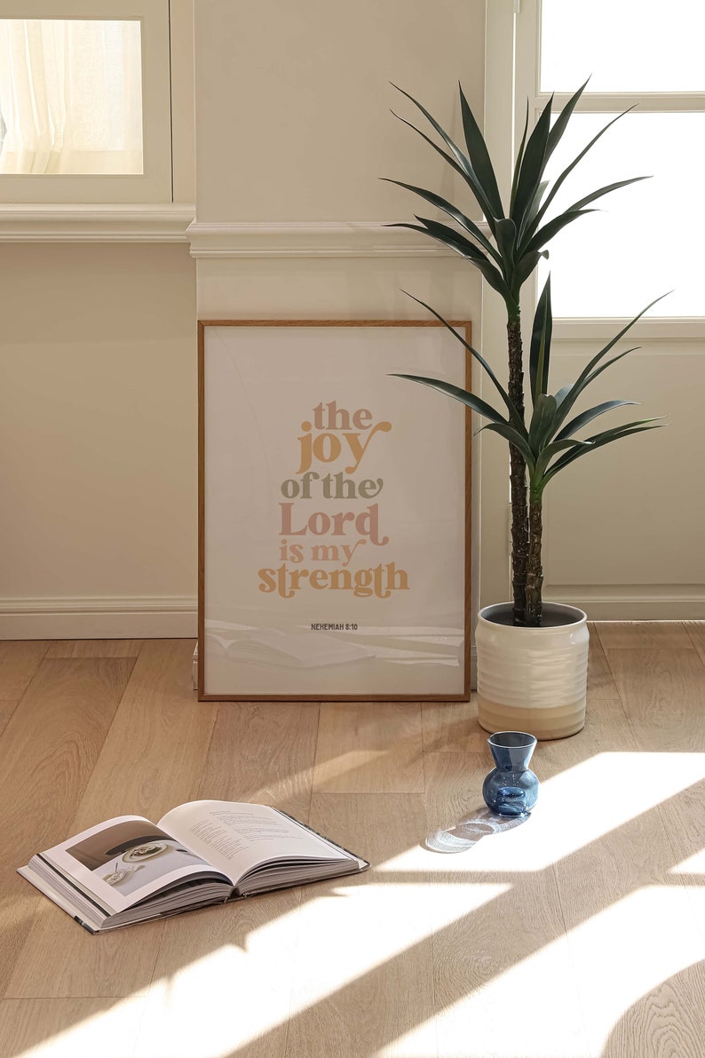 May include: A framed print with the text "the joy of the Lord is my strength" in a warm color palette. A potted plant, an open book, and a small blue vase are also visible on the wooden floor.