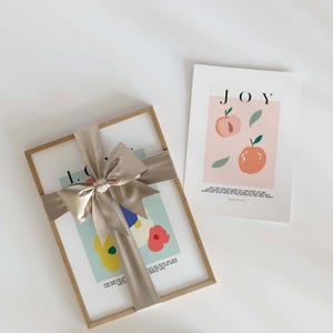 Puede incluir: Dos impresiones art&iacute;sticas enmarcadas. Una impresi&oacute;n est&aacute; envuelta con una cinta de sat&eacute;n beige atada con un lazo. La impresi&oacute;n enmarcada tiene la palabra "LOVE" y arte floral colorido. La segunda impresi&oacute;n tiene la palabra "JOY" e ilustraciones de melocotones.