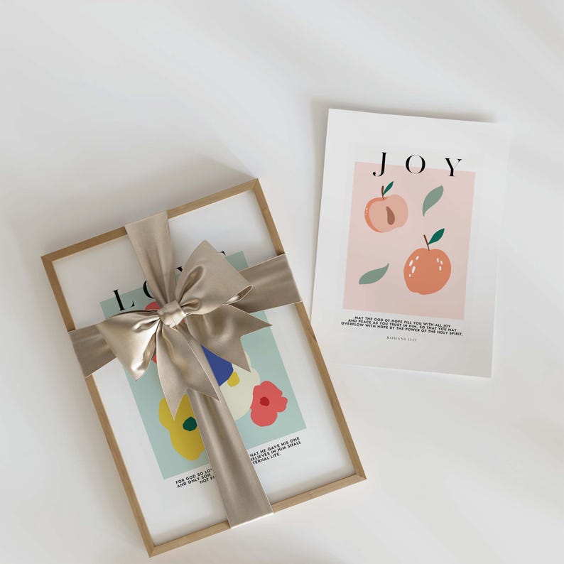 Puede incluir: Dos impresiones art&iacute;sticas enmarcadas. Una impresi&oacute;n est&aacute; envuelta con una cinta de sat&eacute;n beige atada con un lazo. La impresi&oacute;n enmarcada tiene la palabra "LOVE" y arte floral colorido. La segunda impresi&oacute;n tiene la palabra "JOY" e ilustraciones de melocotones.
