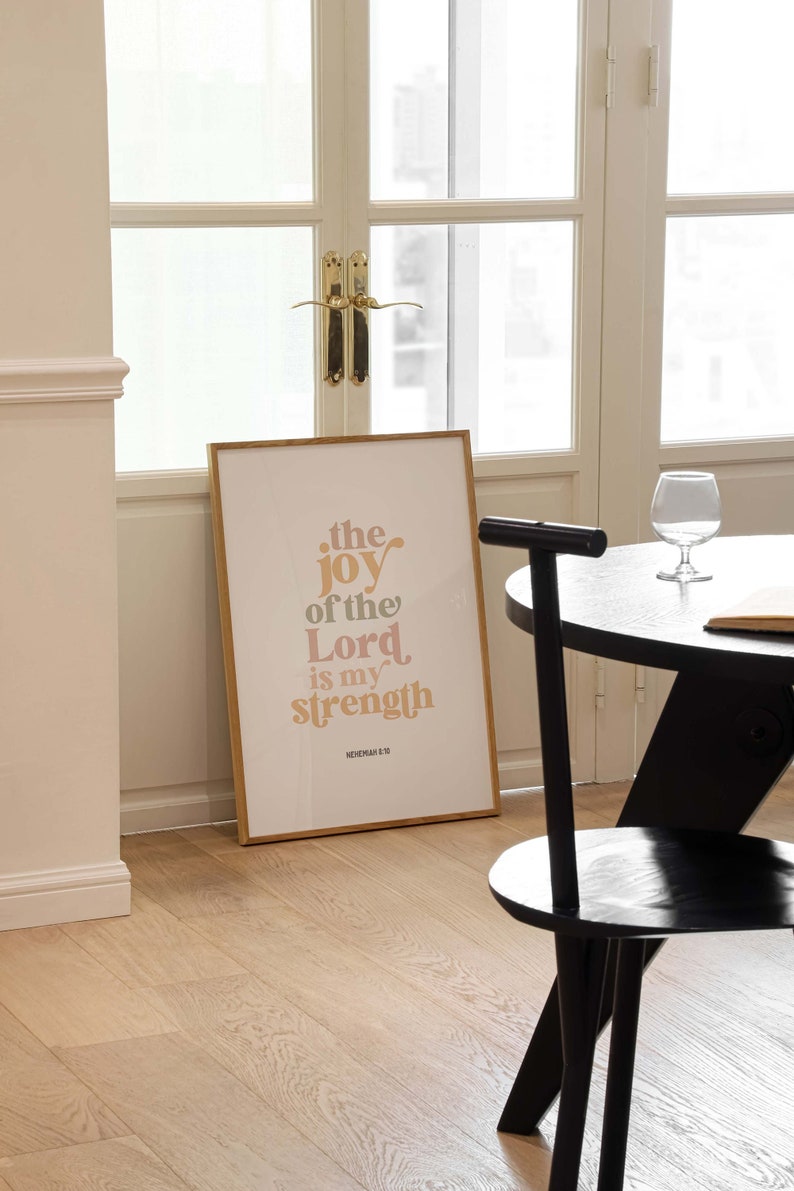 May include: A framed print with the text "the joy of the Lord is my strength" in a warm color palette. The print is displayed in a room with a black chair, a round table, and a glass of water.
