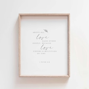 May include: A framed print with a light wood frame. The white background features elegant black script that reads "ABOVE ALL love EACH OTHER DEEPLY. BECAUSE love COVERS A MULTITUDE OF SINS. I PETER 4:8". A small floral sprig is at the top.