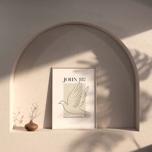 May include: A minimalist art print featuring a white dove illustration and the text "JOHN 8:12" in black. The print is framed in a light wood frame and is displayed on a shelf against a beige wall.