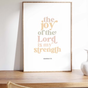 May include: A framed print with the text "the joy of the Lord is my strength" in pastel colors. The print is displayed on a wooden desk with a woven chair in front of it. The text is from Nehemiah 8:10.