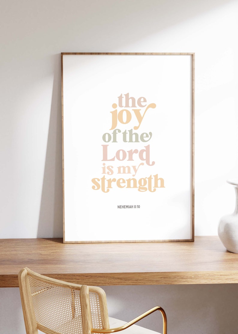 May include: A framed print with the text "the joy of the Lord is my strength" in pastel colors. The print is displayed on a wooden desk with a woven chair in front of it. The text is from Nehemiah 8:10.