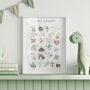 May include: A framed "Bible Alphabet" print with colorful illustrations of biblical themes. A green dinosaur figurine and a cactus-shaped decoration sit on a shelf with books. A garland of green triangles hangs above.