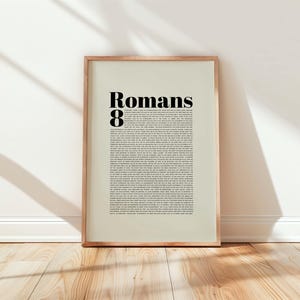 Puede incluir: Un cuadro enmarcado con el texto "Romans 8" en letras negras en negrita en la parte superior, seguido de texto más pequeño. El marco es de color marrón claro y el fondo es de color beige claro. El cuadro está apoyado contra una pared blanca sobre un suelo de madera.