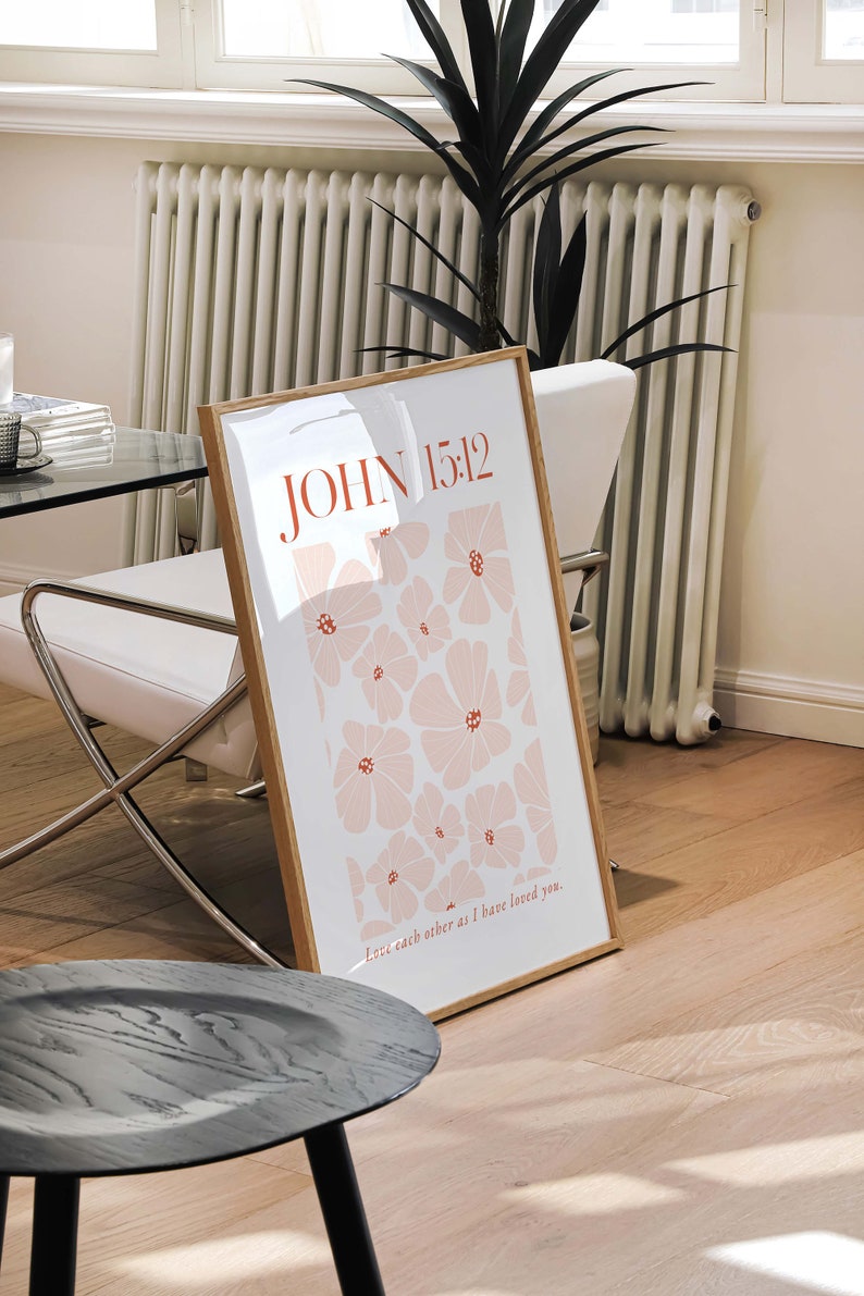 May include: A framed print with a white background and a pattern of pink flowers. The text "JOHN 15:12" is in red at the top of the print. The text "Love each other as I have loved you." is in red at the bottom of the print.