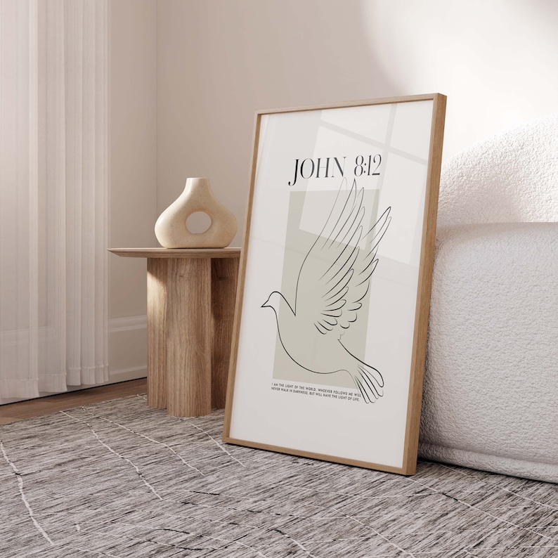 May include: A minimalist art print featuring a dove in flight and the text "JOHN 8:12" in black. The dove is outlined in black against a light beige background. The text "I am the light of the world, whoever follows me will never walk in darkness, but will have the light of life." is printed in black at the bottom of the print.