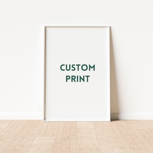 Puede incluir: Una impresión enmarcada en blanco con las palabras "CUSTOM PRINT" en letras verde oscuro. La impresión está apoyada contra una pared blanca sobre un suelo de madera clara.