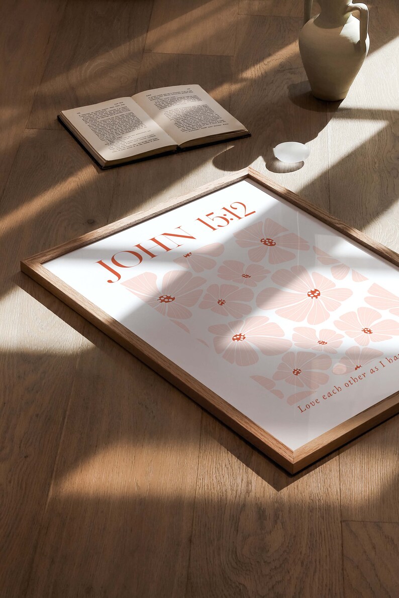 May include: A framed print with a white background and a pattern of pink flowers. The text "JOHN 15:12" is in orange at the top of the print. The text "Love each other as I have loved you" is in orange at the bottom of the print.