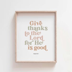 Könnte beinhalten: Ein gerahmter Druck mit beigefarbenem Hintergrund und einem hellbraunen Rahmen. Der Druck zeigt den Text "Give thanks to the Lord, for He is good. Psalm 107:1" in einer stilisierten Schriftart mit Grün-, Rosa- und Brauntönen.