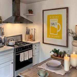 May include: A kitchen with a white stove and a framed print on the wall that says "Be the salt of the earth" with a yellow illustration of a salt shaker. There is a wooden table with a plate and a cup on it.