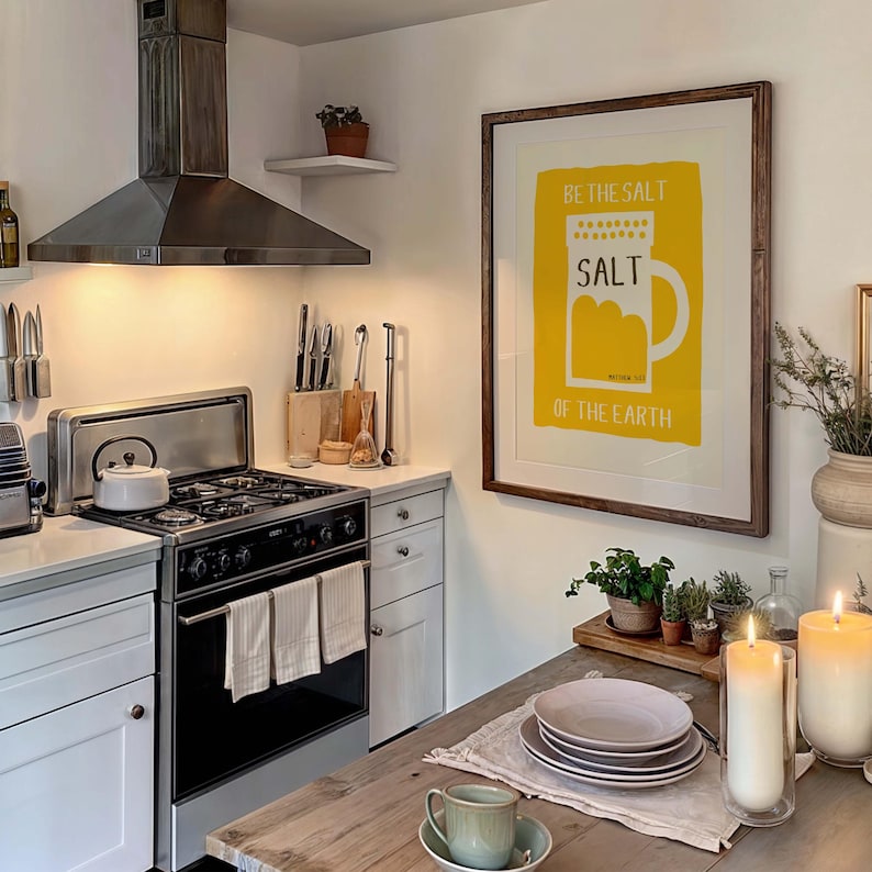 May include: A kitchen with a white stove and a framed print on the wall that says "Be the salt of the earth" with a yellow illustration of a salt shaker. There is a wooden table with a plate and a cup on it.