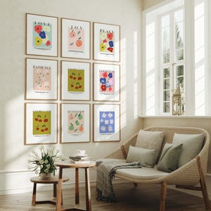 Puede incluir: Nueve impresiones art&iacute;sticas enmarcadas con ilustraciones bot&aacute;nicas y palabras inspiradoras como "Amor" y "Alegr&iacute;a" cuelgan de una pared. Las impresiones presentan flores coloridas, frutas y dise&ntilde;os sencillos. Un sof&aacute; de mimbre con cojines y una peque&ntilde;a mesa de madera est&aacute;n frente a una ventana.