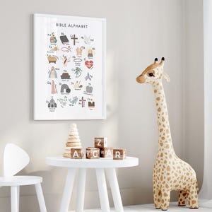 May include: A framed "Bible Alphabet" print hangs on a wall, featuring colorful illustrations of biblical symbols and letters. A giraffe toy with brown spots stands nearby. A white table holds wooden blocks and a stack of wooden rings.