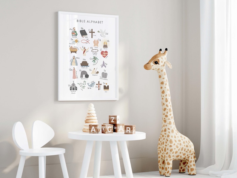 May include: A framed "Bible Alphabet" print hangs on a wall, featuring colorful illustrations of biblical symbols and letters. A giraffe toy with brown spots stands nearby. A white table holds wooden blocks and a stack of wooden rings.