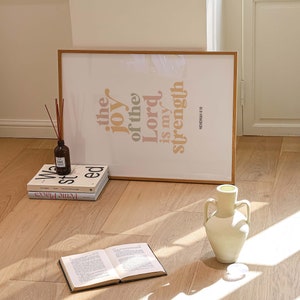 May include: A framed print with the text "the joy of the Lord is my strength" in orange and peach hues. A stack of books, a vase, and an open book are on the wooden floor. A diffuser bottle with reeds is also present.