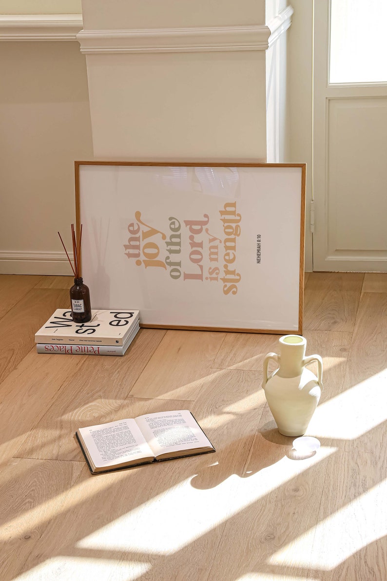 May include: A framed print with the text "the joy of the Lord is my strength" in orange and peach hues. A stack of books, a vase, and an open book are on the wooden floor. A diffuser bottle with reeds is also present.