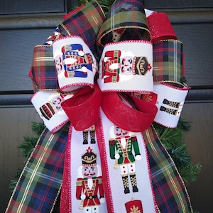 May include: A Christmas bow with red, green, and blue plaid ribbon and white ribbon with a repeating pattern of nutcracker figures.