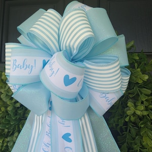 May include: A blue and white striped bow with a blue glitter ribbon and a white ribbon with the text "Baby!" and a blue heart.