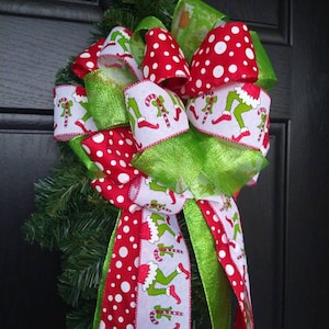 May include: A festive Christmas bow featuring red and white polka dot ribbon, green ribbon, and ribbon printed with a whimsical green character holding a candy cane. The bow is attached to a small evergreen base.